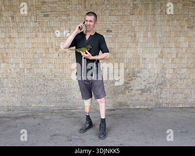 A senior man with a prosthetic left leg is talking on a smartphone and holding a small green parrot Stock Photo