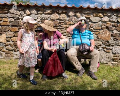 A family resting on a wooden bench against a stone wall on a sunny day. A man is drinking from a bottle, while a woman and children sit nearby Stock Photo