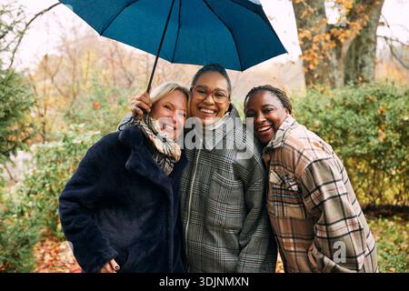 Portrait of smiling multiracial women leaning on female friend while standing under umbrella in rain Stock Photo