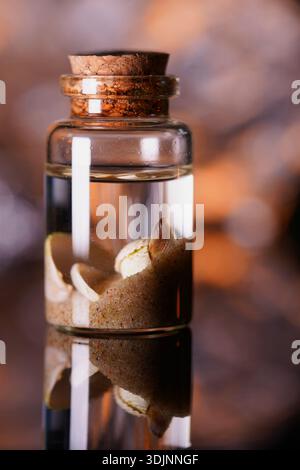 Small glass apothecary bottle with cork stopper containing sea shells and sand in water, reflected on dark glossy surface Stock Photo