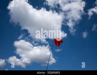 A single red heart-shaped foil balloon floating against a bright blue sky with fluffy white clouds on a sunny day Stock Photo