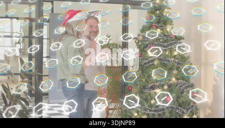 Hugging senior couple wearing sweaters and Santa hat, sharing embrace in lounge with Christmas tree Stock Photo