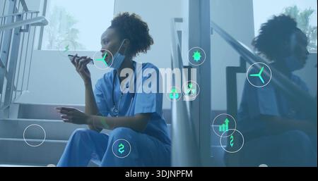 Sitting nurse in blue scrubs speaking into phone on stairwell, with surgical mask lowered, AR icons Stock Photo