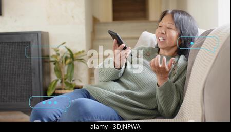 Reclining senior in green sweater holding black phone, gesturing on sofa at home, with chat bubbles Stock Photo