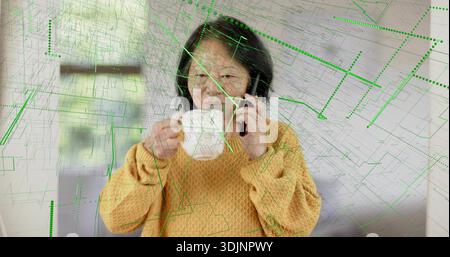 Talking senior Chinese woman wearing mustard sweater holding white mug on phone at home, green-grid Stock Photo