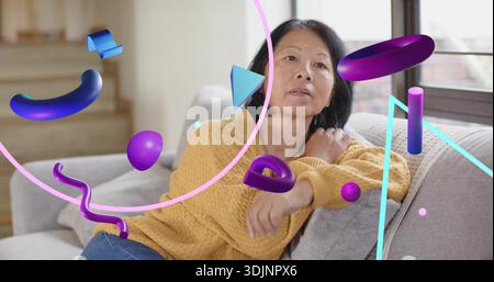 Reclining senior Chinese wearing mustard sweater and blue jeans on sofa, purple 3D shapes floating Stock Photo