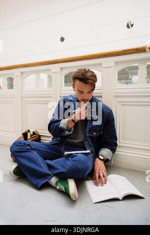Focused young male student studying while sitting cross-legged on ground in corridor at college Stock Photo