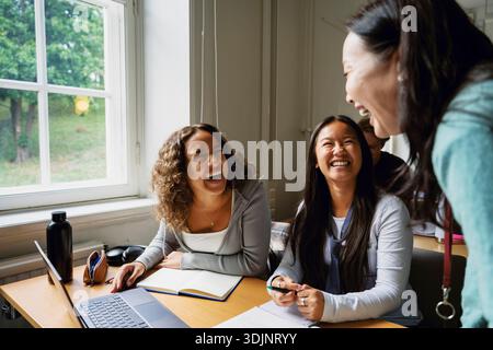 Female students laughing with teacher while sitting in classroom at college Stock Photo