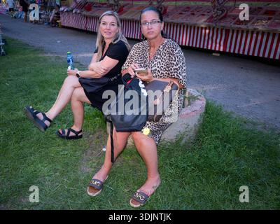 Two 40s women sitting on a concrete edge at a funfair, one looking at her smartphone Stock Photo