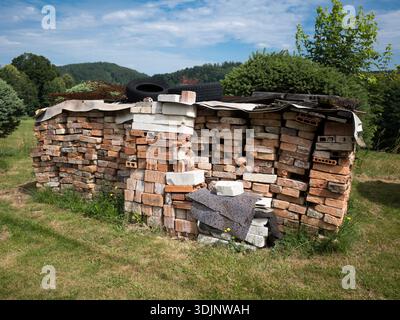 A large stack of old red and white bricks covered with a tarp and tires, standing in a grassy field with green hills and trees in the background. Stock Photo