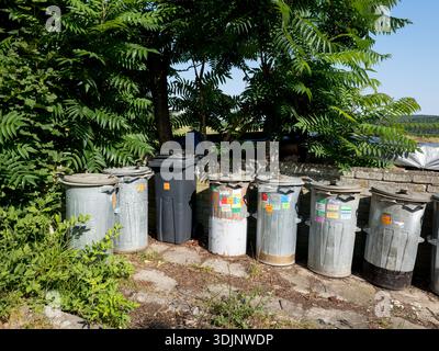 A row of weathered old metal garbage bins and one black plastic bin standing on a stone pavement against a stone wall and green trees in a backyard. Stock Photo