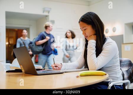 Young female student leaning on elbow writing in book while sitting at table Stock Photo
