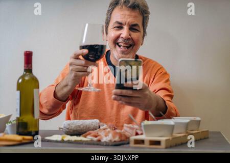 A happy middle-aged man enjoys a relaxed evening at home, holding a glass of red wine and using his smartphone while sitting at a table with an Italia Stock Photo