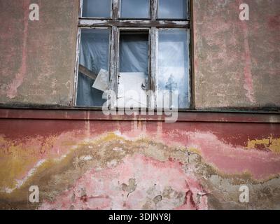 Detailed view of a weathered red and yellow facade with a large wooden window on an old abandoned building. Peeling paint and decaying architecture Stock Photo