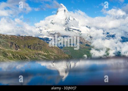 View of majestic Matterhorn peak piercing the sky, surrounded by fluffy clouds and reflected in serene waters, Zermatt, Valais, Switzerland. Stock Photo