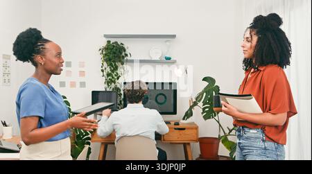 Collaborative Office Scene With Two Women Talking As Colleague Works At Desk In Modern Workspace Stock Photo