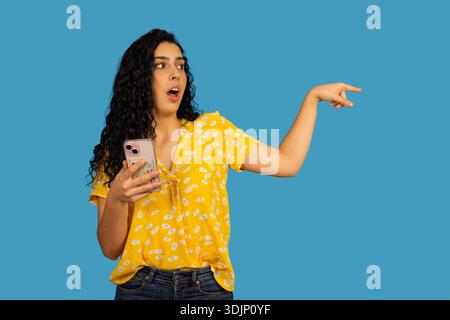 Woman feeling surprised and pointing, holding a smartphone with a blank expression Stock Photo