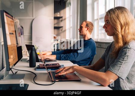 Blond female programmer coding on laptop and computer while sitting at desk in startup office Stock Photo