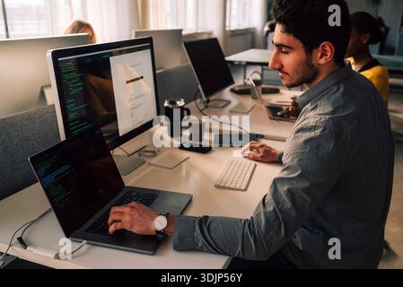 Young male programmer typing on laptop while sitting at desk in tech office Stock Photo