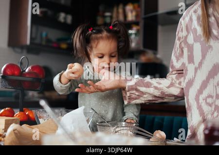 A child reaches out to an adult in the kitchen while holding an egg. They are preparing to bake with various ingredients and utensils on the table in Stock Photo