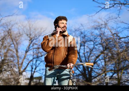 Stylish young man communicating on a cell phone while pushing his bicycle through an urban park in winter Stock Photo
