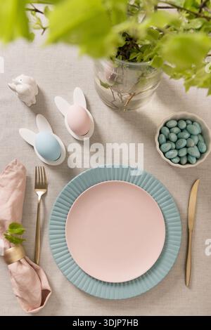 Easter flat lay with pastel plates, decorative eggs, bunny holders, spring branches and sweets on linen tablecloth. Cozy holiday table decor, modern l Stock Photo