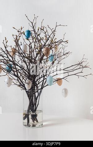 Decorative Easter branches with hanging pastel and speckled eggs in glass vase on white background. Spring holiday decoration, minimal composition, ve Stock Photo