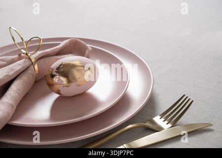 Elegant Easter table setting with pastel pink plates, decorative egg with gold leaf and golden cutlery on light linen tablecloth. Luxury spring holida Stock Photo
