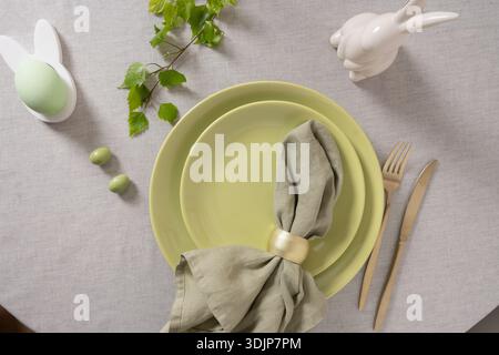 Easter flat lay with green plates, bunny shaped egg holder, linen napkin, golden cutlery and spring greenery on neutral background. Modern seasonal ta Stock Photo