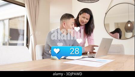 Reviewing couple in pink and gray sweaters using laptop at dining table, with blue like graphic Stock Photo