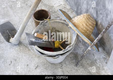 Messy construction site featuring tool bucket trowel hoe and hat resting on rough concrete floor showing busy work in progress atmosphere Stock Photo
