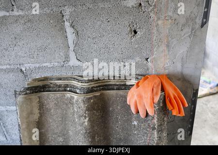 Close up of heavy duty orange rubber gloves on gray corrugated concrete roof tile against rough unfinished cinder block wall showing grit and construc Stock Photo