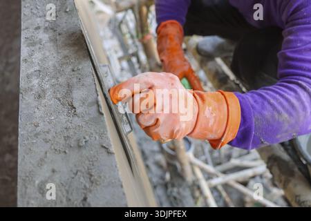 Closeup of meticulous male construction worker hand wearing orange protective glove carefully plastering wet grey cement or concrete onto wall with fo Stock Photo