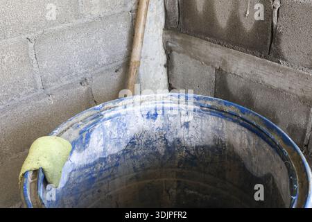 Dirty blue plastic barrel standing near grey concrete block wall corner with old wooden broom handle and yellow sponge rim evoking feeling of abandone Stock Photo