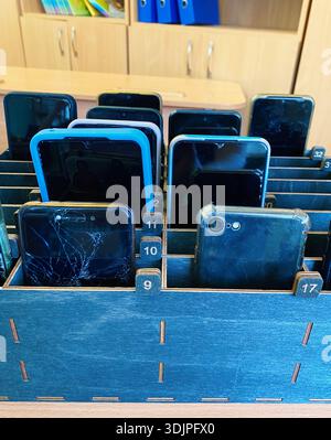 Wooden organizer holds several smartphones with cracked screens and different colors, arranged in numbered slots on a desk in a classroom environment Stock Photo