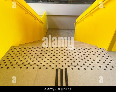 Bright yellow stairs descend with tactile paving for safety, complemented by handrails and a sleek concrete floor in a contemporary interior setting Stock Photo