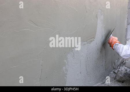 Builder plastering wall with trowel carefully working on construction site grey cement surface smooth finish concrete texture background creates focus Stock Photo
