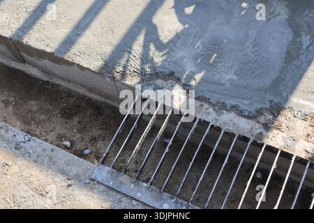 Close up shot of dirty metal grate drain cover on concrete floor outdoors with shadow patterns casting over surface creating rough and gritty texture Stock Photo