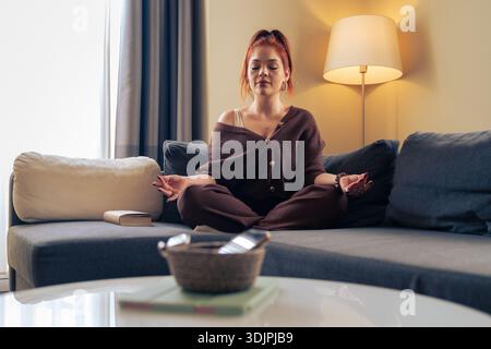 Young woman meditating on a sofa at home with a calm expression, while smartphone and tablet lie on the table nearby. Concept of mindfulness, wellness Stock Photo