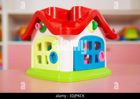 Brightly colored plastic toy house featuring a red roof and various doors, positioned on a pink cabinet with additional toys visible in the background Stock Photo