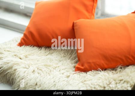 Two orange pillows rest on a fluffy white rug beside a window, enhancing the comfort of the indoor space with a warm color contrast Stock Photo