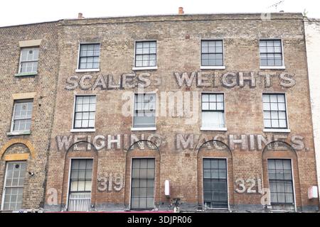 Faded painted sign reading ‘Scales Weights & Weighing Machines’ on a Victorian brick building facade in East London, numbered 319-321. Stock Photo