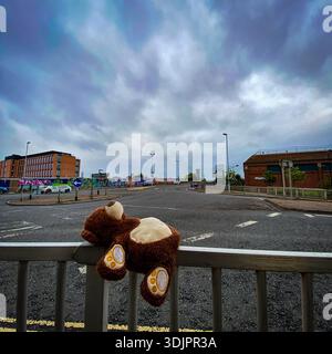 Lonely teddy bear draped over a barrier rail on an empty urban roundabout near modern buildings and graffiti murals under a cloudy sky in England Stock Photo
