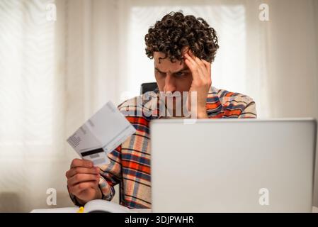 Stressed man using laptop and credit card to pay his financial bills online while sitting at home. Stock Photo