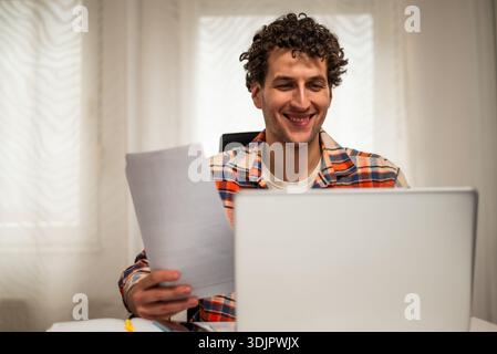 Cheerful young freelancer using laptop and checking documents while working at his cozy home. Stock Photo