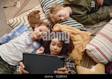 Multiethnic Generation Alpha Children Lying on Pillows Sharing Tablet Experience Stock Photo