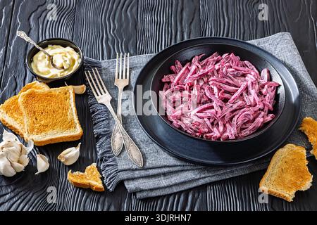 Traditional Eastern European beetroot salad with garlic and mayonnaise in a black bowl on a dark wooden table with toasted bread and forks, horizontal Stock Photo