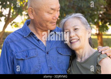 Senior Asian couple posing in garden, man in blue denim shirt, woman in olive-green top, smiling Stock Photo