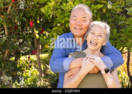 Senior Asian couple embracing, laughing, standing in sunlit garden wearing denim shirt, smartwatch Stock Photo