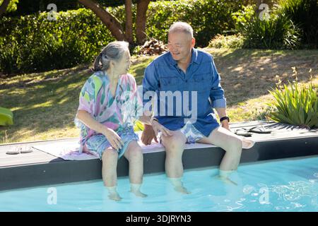 Asian senior couple sitting on pool edge, dipping feet, chatting with towel, sandals, shades nearby Stock Photo